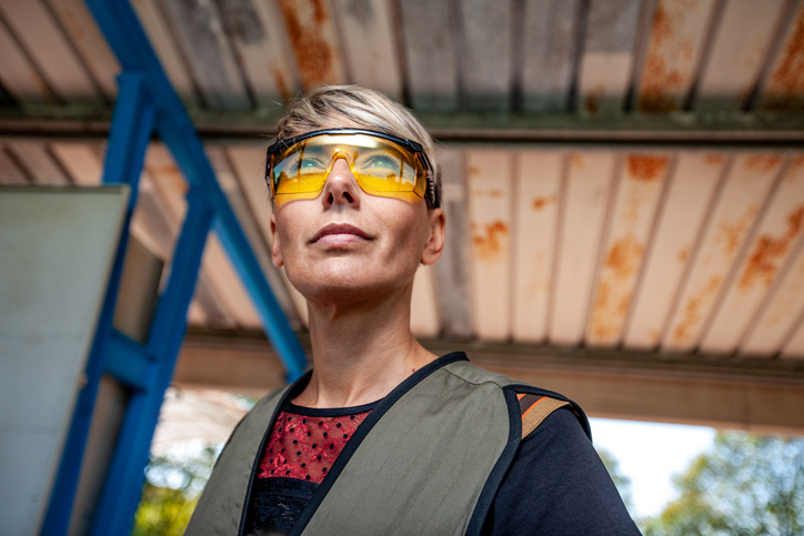 A middle aged women, waist up, with yellow safety glasses looks down range at the gun range.