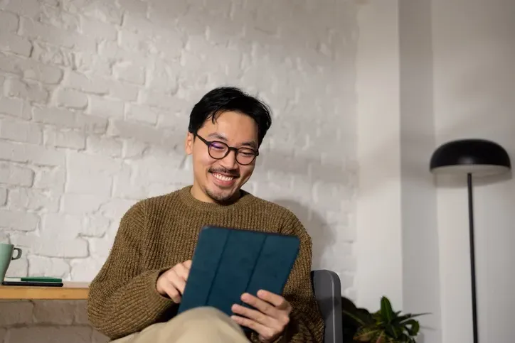 An Asian guy is looking at his iPad and smiling while sitting on an office chair.