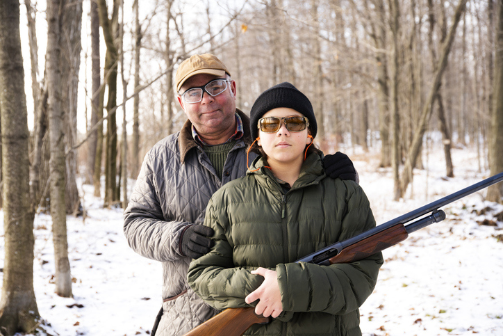 A father stands behind his son in the woods. The son standing in front of the dad holds a shotgun pointed safely at the woods.