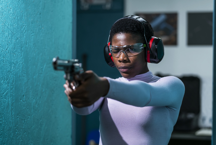 A close-up shot of a woman in safety glasses and earplugs is aiming a handgun down range at a target at a gun range.