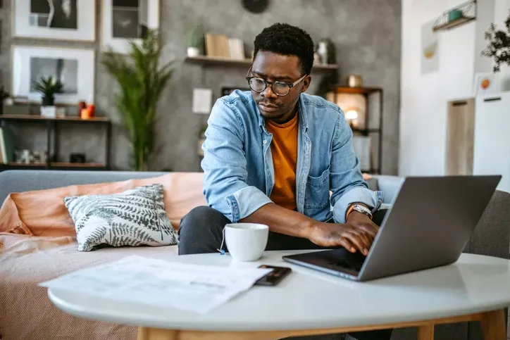 A black man is working on his computer in his living room while looking at pieces of paper on a coffee table next to him.