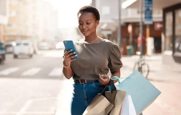 A black women smiling and holding shopping bags while looking at her phone standing on the sidewalk.