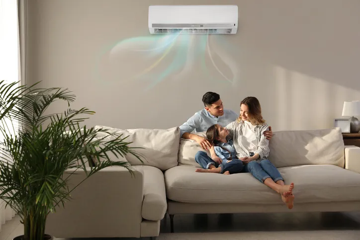 A family is sitting on a couch with an AC unit above them on the wall cooling the living room area.