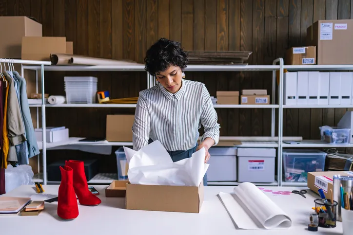 A woman in an office, standing over a table while she opens a box with tissue paper. A pair of red high heels are also on the table.