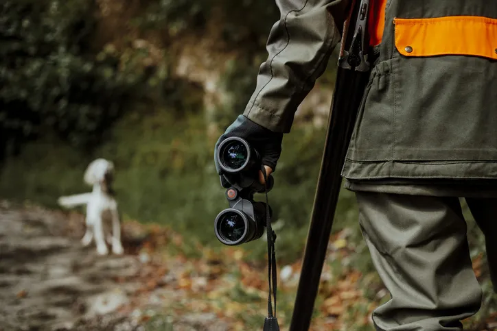 A hunter holding binoculars and a rifle, with a dog in the background, during a hunting expedition.