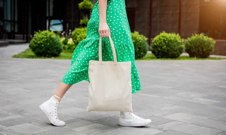 A woman walking in a green dress is pictured from the waist-down carrying a reusable shopping bag.