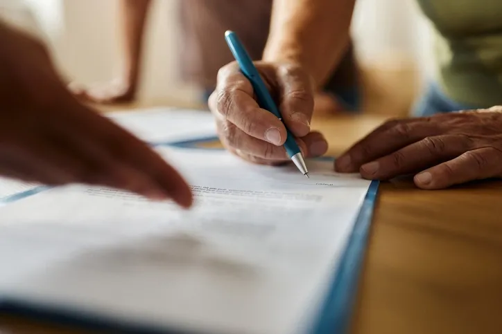 A close-up of a person signing a contract while another person's hand points where to sign.
