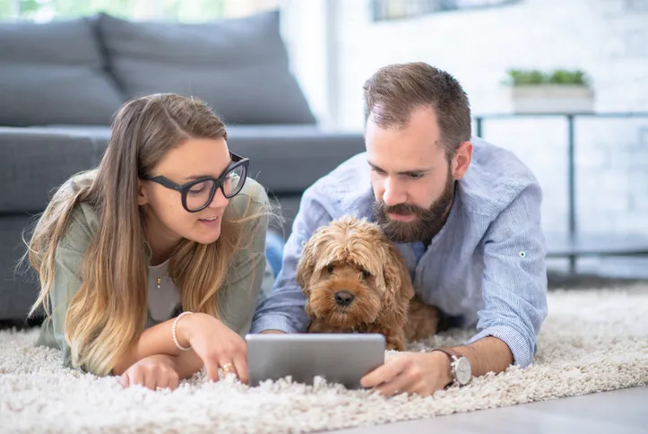 A man and woman are laying on their stomachs on the living room floor with their dog looking at an iPad while smiling and pointing at it.