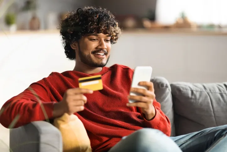A young man with curly hair is sitting on a couch holding a credit card and computer while smiling.