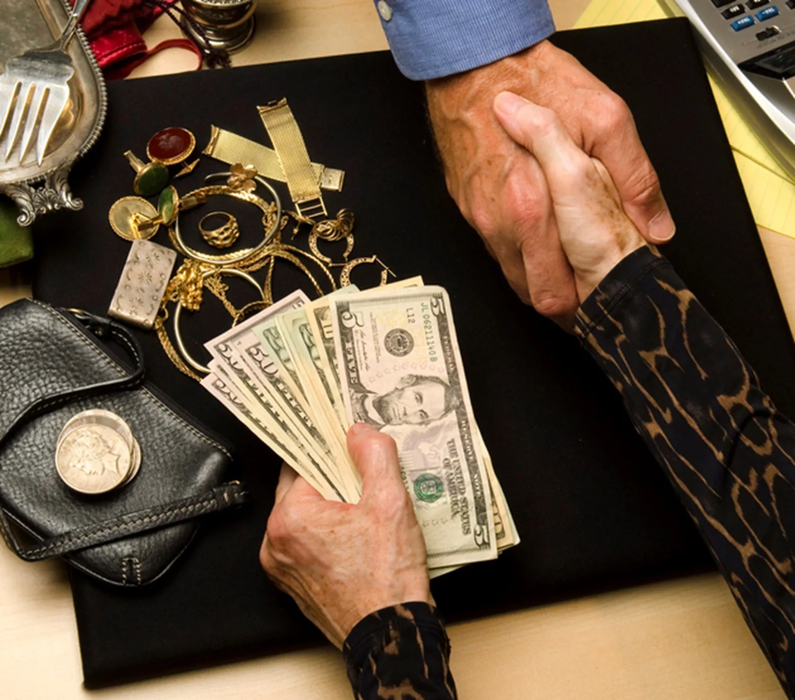 A women holding cash in her left hand is shaking a man's hand with her right hand. There is gold jewelry on the counter. It looks like a sale.