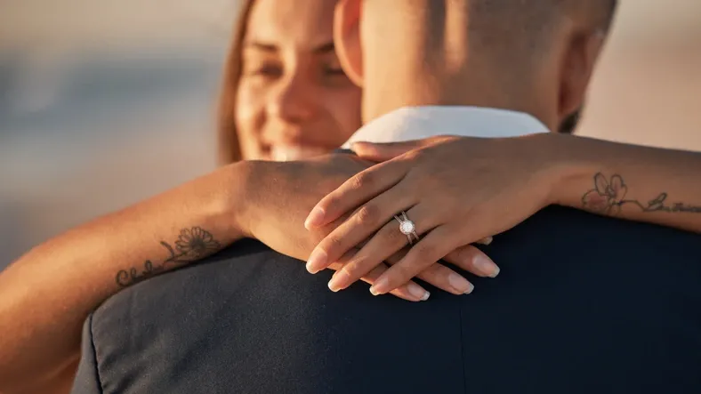 A women with a diamond ring on her ring finger and smiling is hugging a man wearing a suit on a beach with the sun setting.