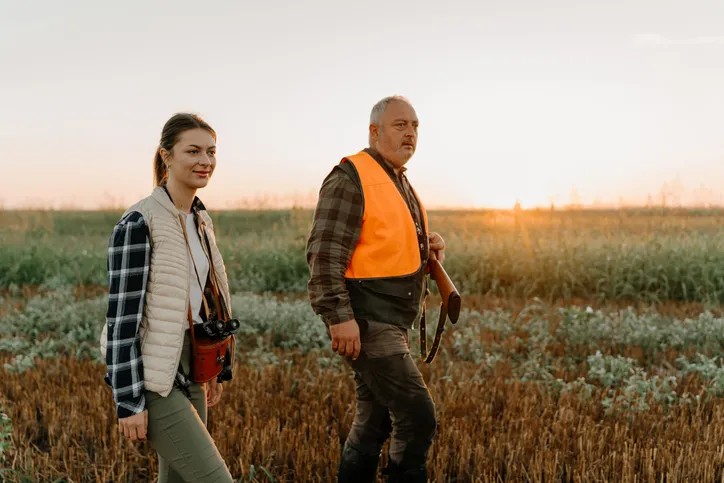 A man and a woman hunter, walking through a field at sunset, with the man carrying a rifle. The woman is equipped with binoculars, indicating a hunting activity.