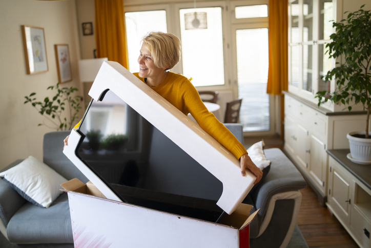 An older woman in her living room is pulling a new TV out of a box while smiling.