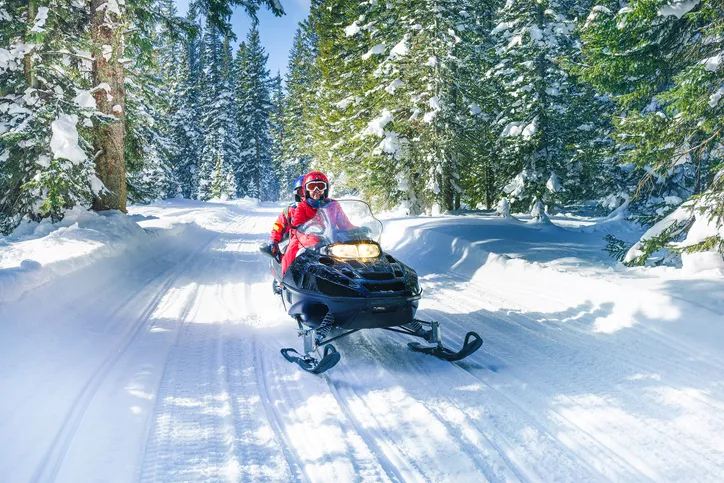Two people are seen snowmobiling down a winter trail with pine trees in the background.