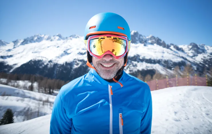 A man on a ski slope is looming directly at the camera with goggles, a helmet and a winter jacket while smiling.