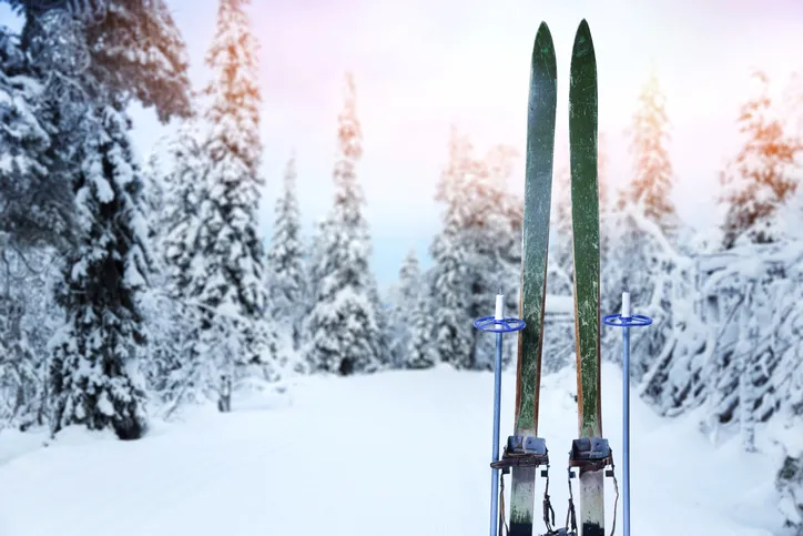 A pair of green skis and ski poles are stuck straight up in the snow with snow and pine trees in the background.