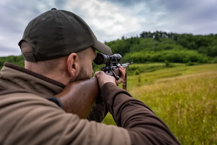 A person aiming a rifle equipped with a scope, while hunting or practicing shooting.