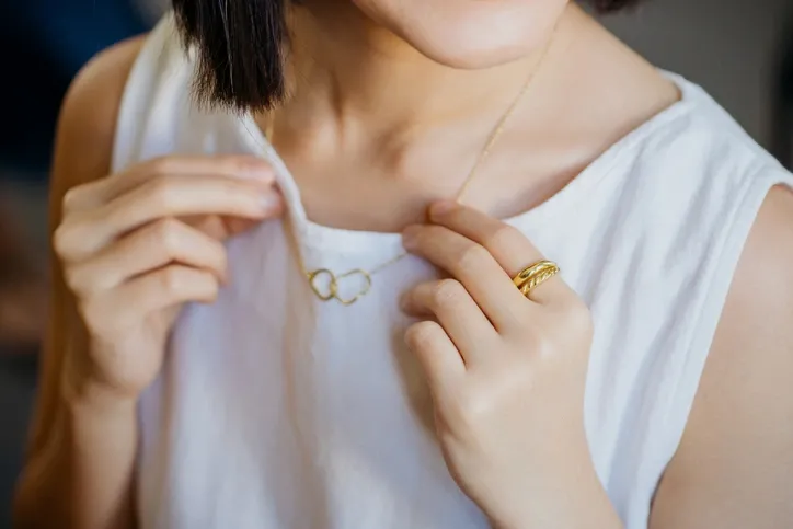 A closeup of a woman holding her gold necklace with hearts in a white tank top. Just her chin to her stomach is in frame.
