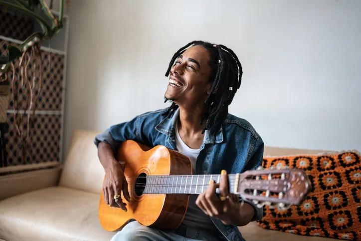 A man is sitting on a couch playing an acoustic guitar while singing and or smiling.