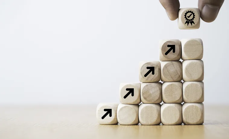 A staircase of cubes with arrows pointing upwards on the top row with the top cube being placed by a hand with a ribbon on it.