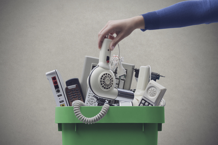 A pile of electronics sits in the trash while a woman places a hairdryer on top.