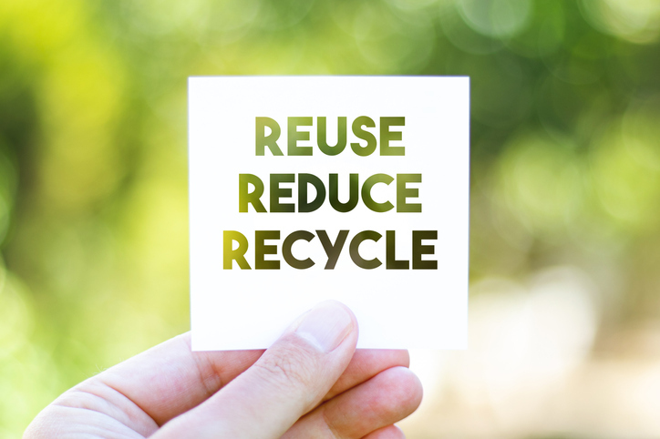 A woman is holding up a white piece of paper with the words, “reuse,” “reduce,’ and “recycle” on it.