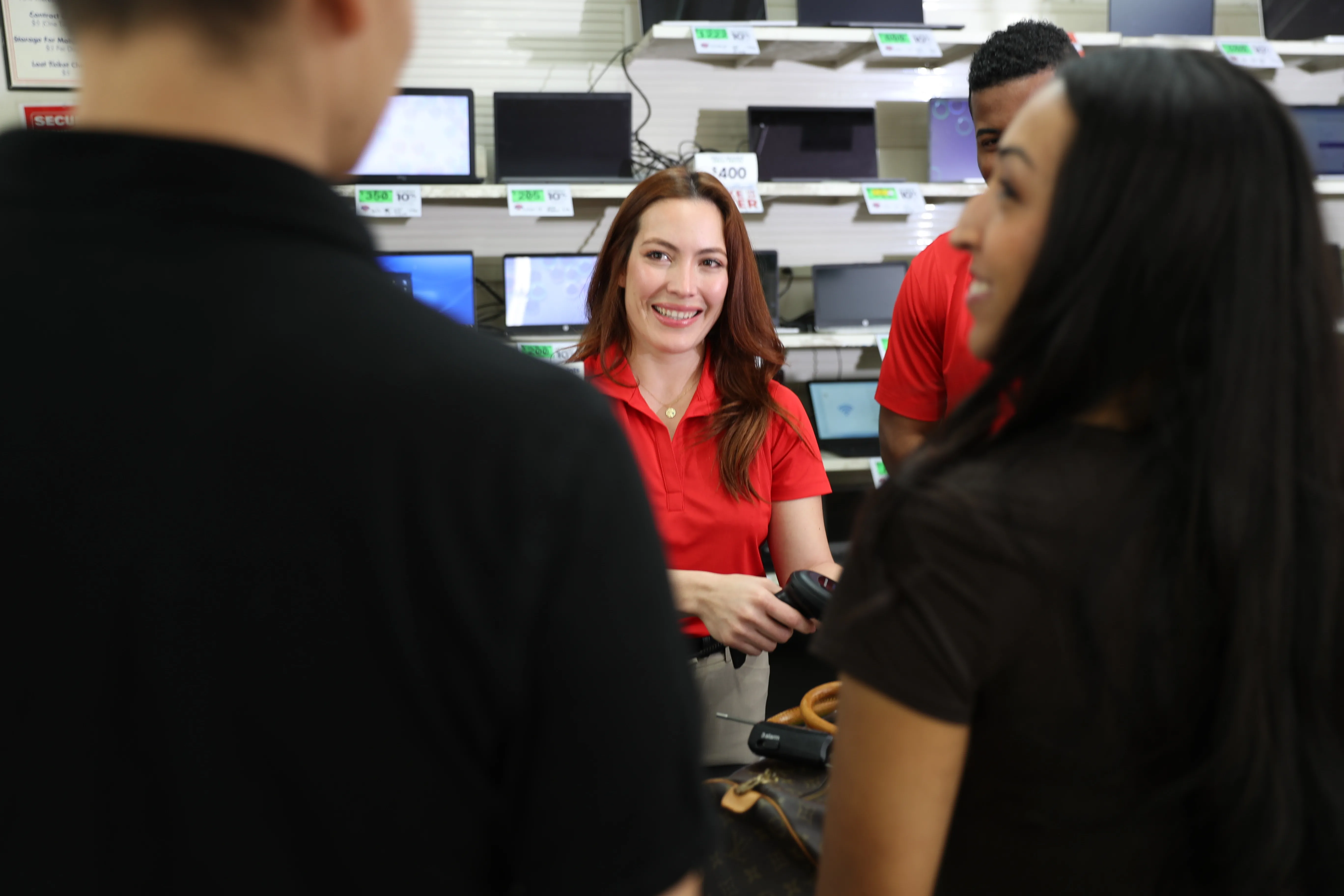A team member in the background is center frame while smiling and checking out items a couple in the foreground have chosen to purchase. They have their backs turned to the camera and are out of focus.