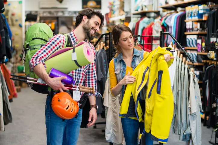 A young couple, a man and women, are looking at sporting equipment in a retail store. The women is holding a yellow rain jacket.