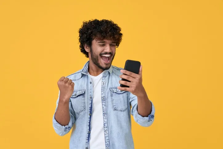 A guy smiling and making a fist while holding his phone in the other hand with a bright yellow background.