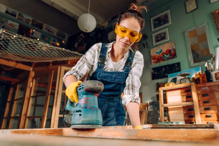 A women is sanding a piece of wood in what looks like her studio. She is wearing proper personal protective equipment (PPE): gloves, safety glasses, and workwear.