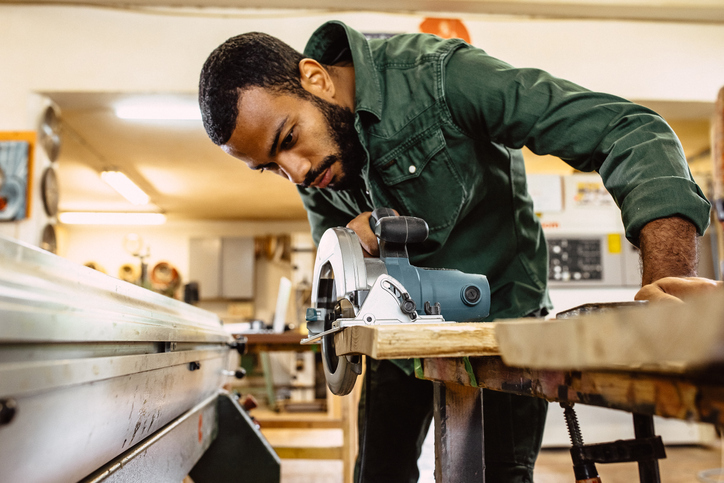 A young black male in a workshop is using a circular saw while cutting a piece of wood.