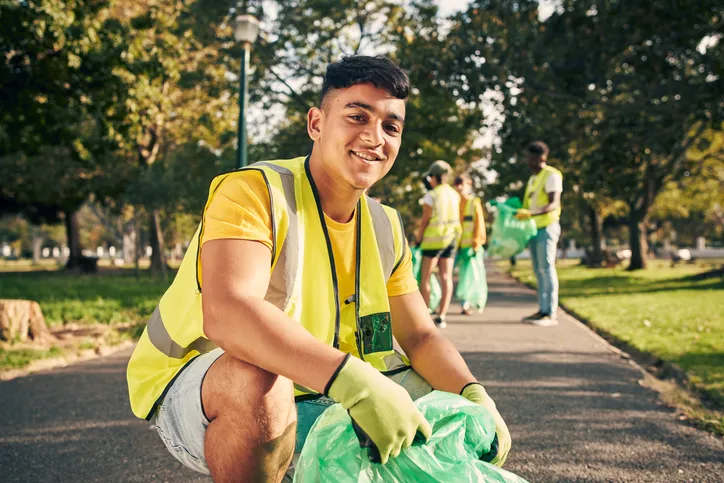 A smiling young man, with a trash bag in his hands, is wearing a safety vest and gloves while picking up trash