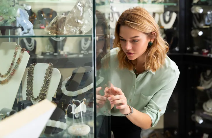 A woman intently looking through a glass display case, examining jewelry, including necklaces and bracelets.