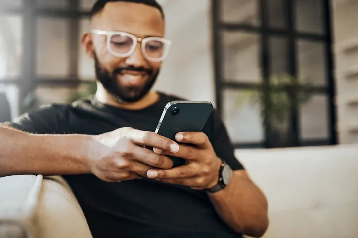 A young professional male is sitting on the couch looking at his phone with translucent glasses while checking his credit score.