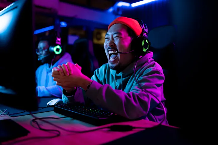 A young man, wearing a headset, laughing and engaged at a gaming setup with multiple monitors.