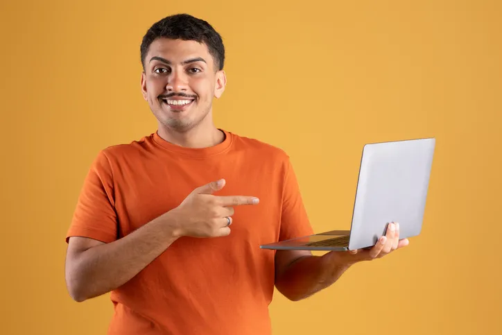 A young guy is holding a laptop while pointing at it and smiling.