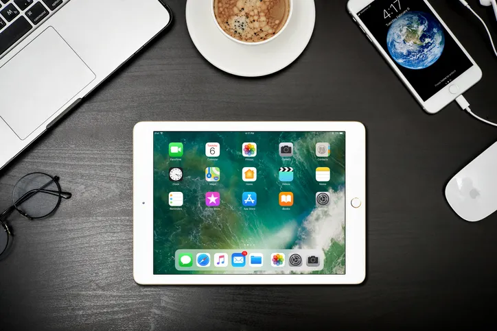 A white iPad is center focus with a laptop, mouse, iPhone, a coffee, and reading glasses surround the edge of the frame.