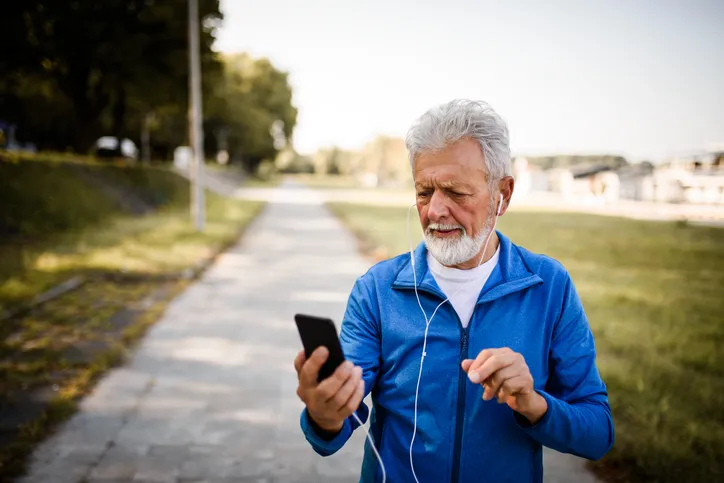 An older man outside is looking at his phone confused. He is in a suburb with headphones in, most likely walking or jogging.
