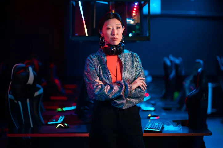 A young Asian gamer is posing with her arms crossed. In the background is a gaming area filled with computers, gaming chairs, screens, neon lights, a table and more.