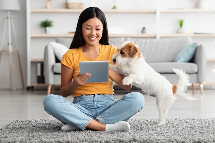 A girl on her iPad and her dog are in a living room while smiling.