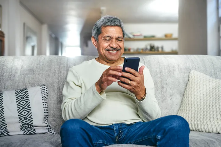 An older man sits on a couch shop on with his phone in hand while smiling.