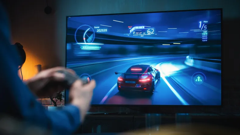 A young man out of frame but with his arms and hands in frame is holding a controller while playing a racing game. A car is drifting on the TV in front of the young man.