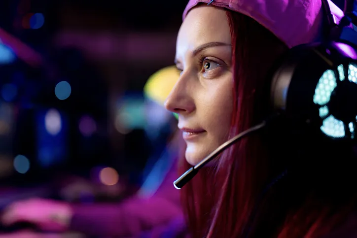 A young women gamer is wearing a headset while looking forward. She is wearing a beanie and smiling slightly.