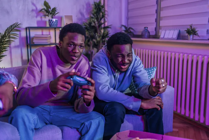 Two young men are playing a video game while sitting on the couch. Something recently happened as they are reacting to whatever is shown on screen.