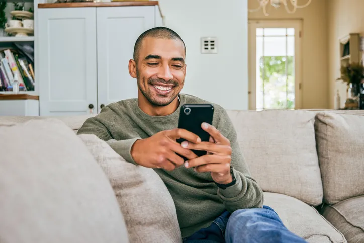 A man sitting on a couch with his phone in hand while smiling.